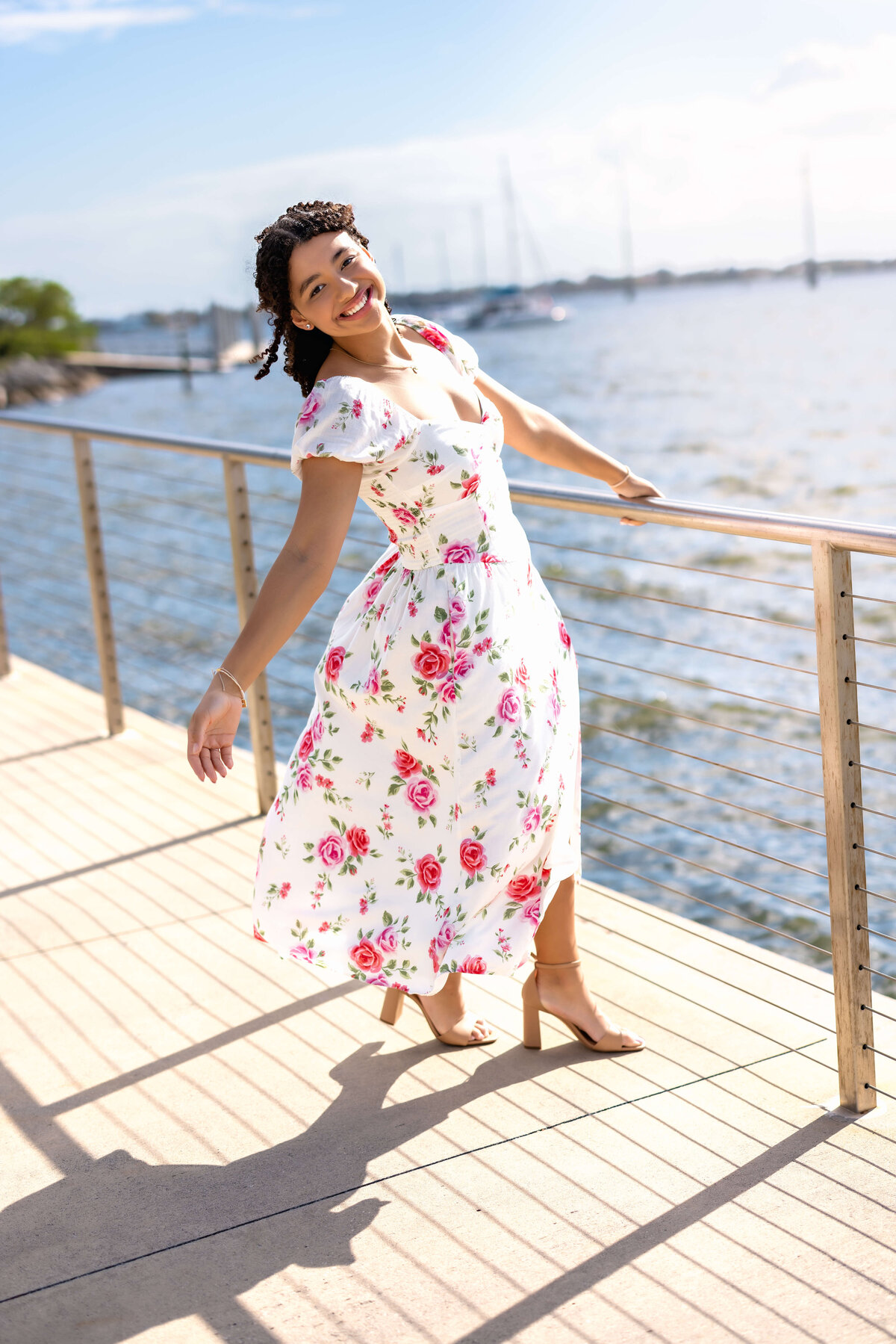 Celebration senior session vibe with a girl leaning outward from a railing, floral dress blowing in the wind as she smiles toward the camrera.