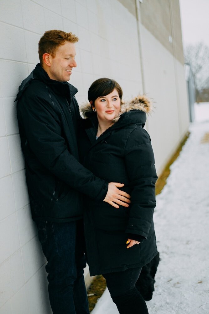 A boyfriend leans against a white wall while embracing his girlfriend