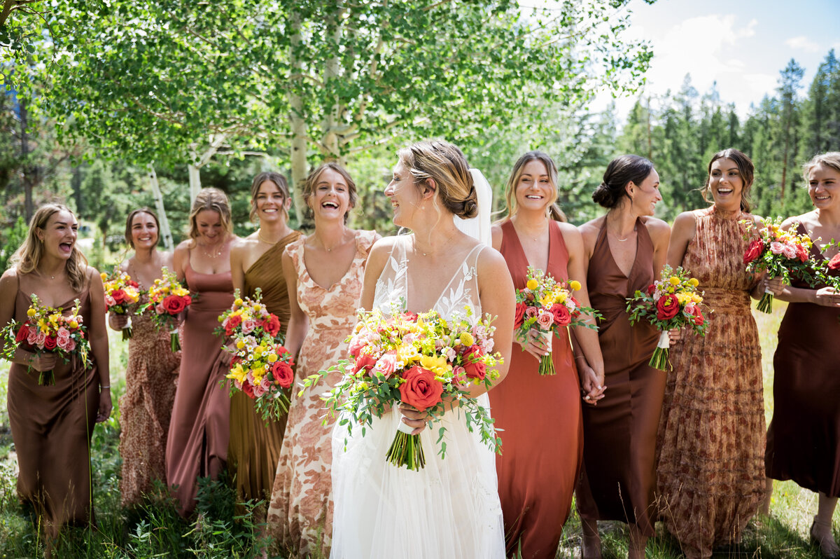 A bride laughs and walks toward the camera with her bridesmaids dressed in a variety of orange dresses.