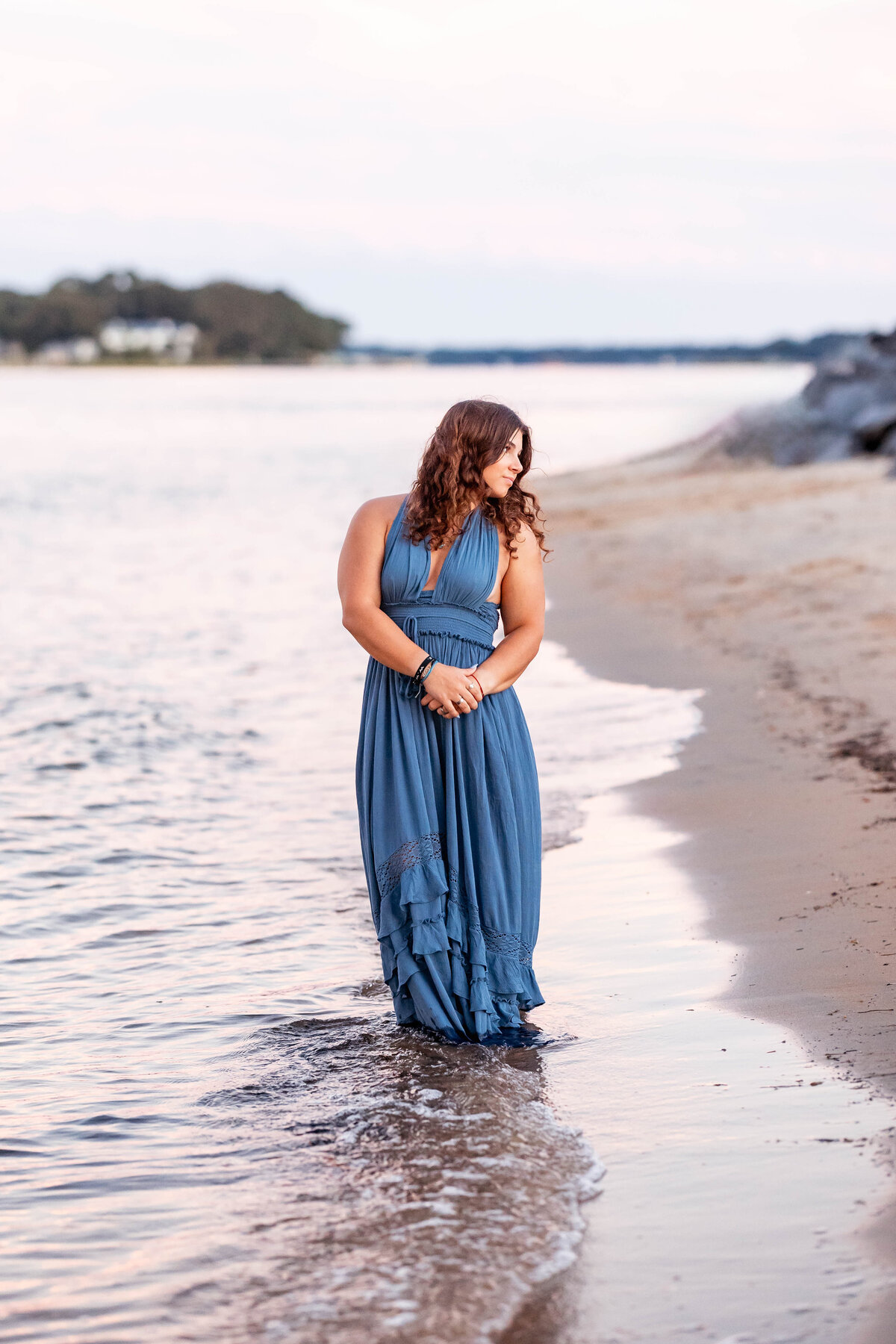 A high school senior in a flowing blue dress stands at the edge of the water in Coastal Virginia, her feet in the surf and her hands gently clasped, framed by soft pastels of sunset and the peaceful rhythm of the waves.