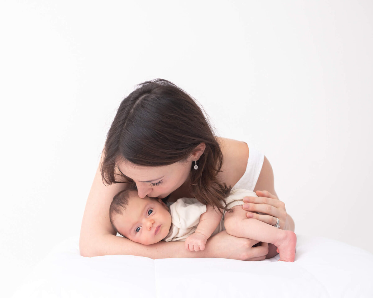 Mother kissing new baby in white themed  professional studio portrait