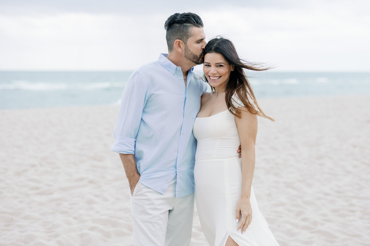Couple enjoying a romantic moment on Miami Beach, with the man kissing the woman on her forehead as they smile, set against a serene ocean backdrop.