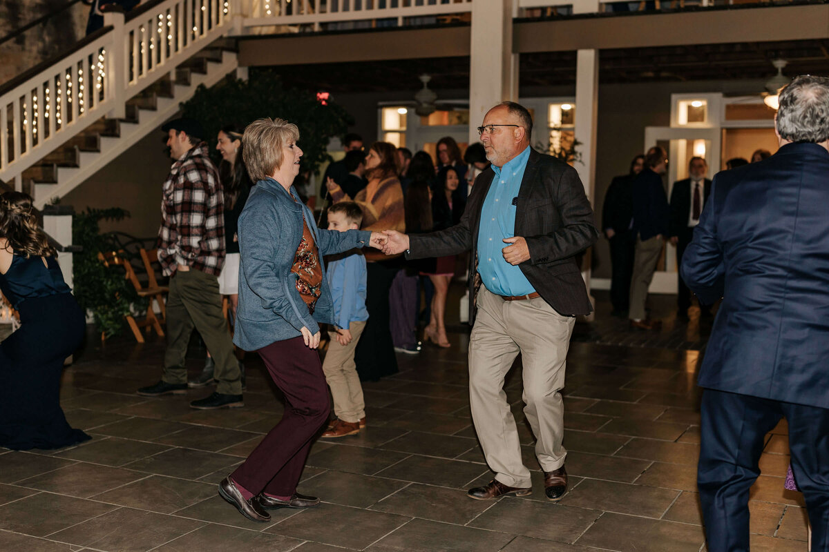 A man and woman dance together in a lively outdoor setting as other people mingle and watch in the background. The atmosphere is festive, with warm lighting and a staircase visible behind the crowd.