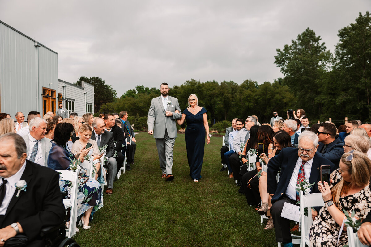 A groom walks down the aisle arm in arm with a woman in a blue dress
