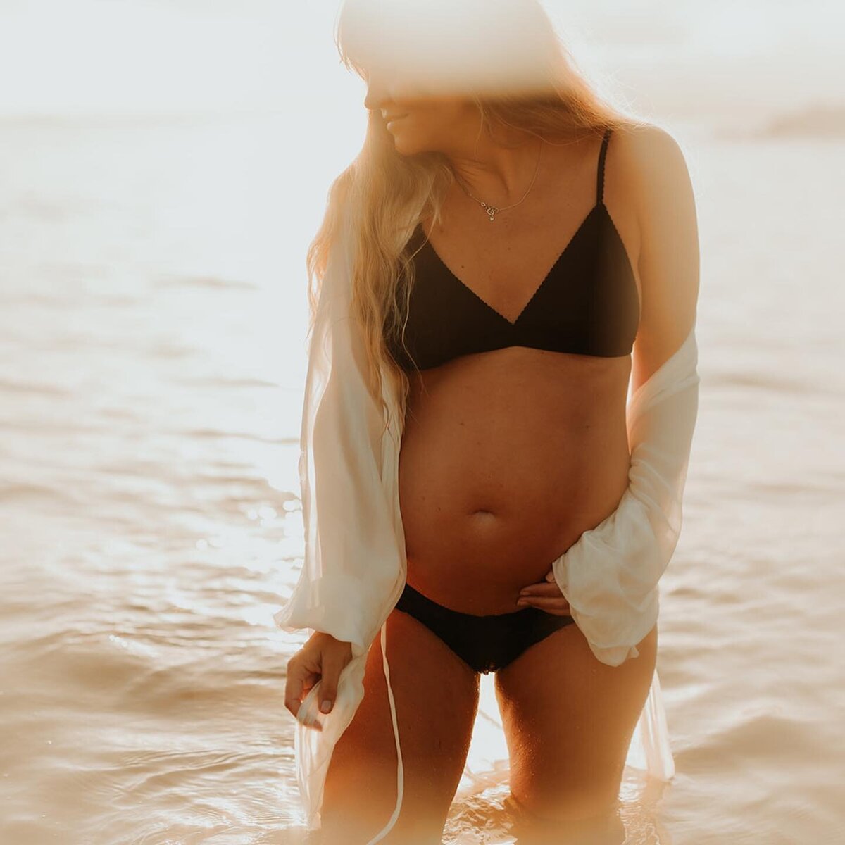 Woman with a black bikini and bares her pregnant belly . her face seems content as she looks down at the water. The Sun is shining behind her