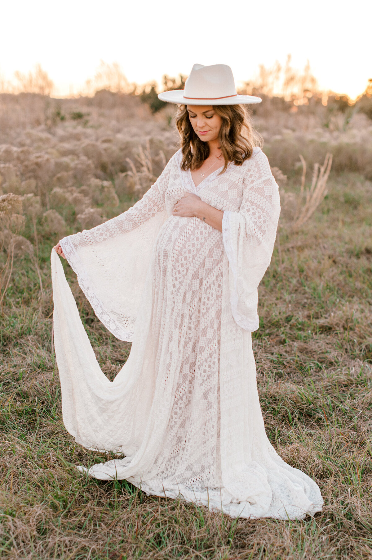 Pregnant mom stands holding her dress and belly at sunset in a field.