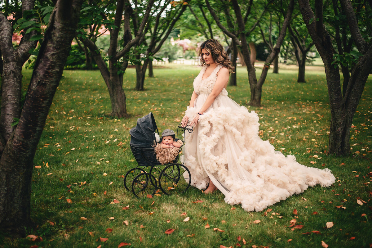 Mom dressed in a lavish cream gown posed pushing her newborn boy in a carriage in Vineland cherry blossom farm by Greater Toronto Newborn Photography session outdoors by Tamara Danielle.