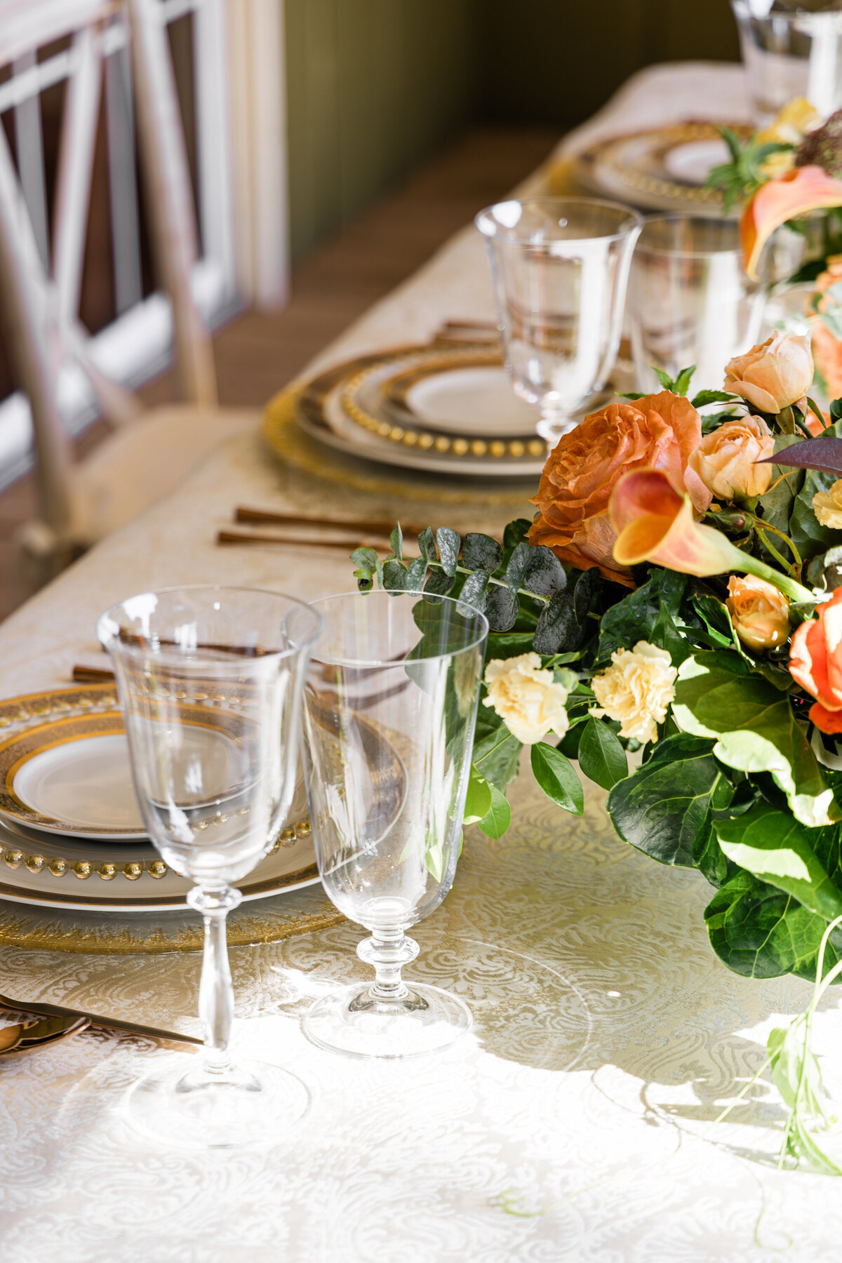 wine and water glasses next to a four plate mixed metals place setting with four piece rose gold flatware set and a orange rose and calla lily centerpiece