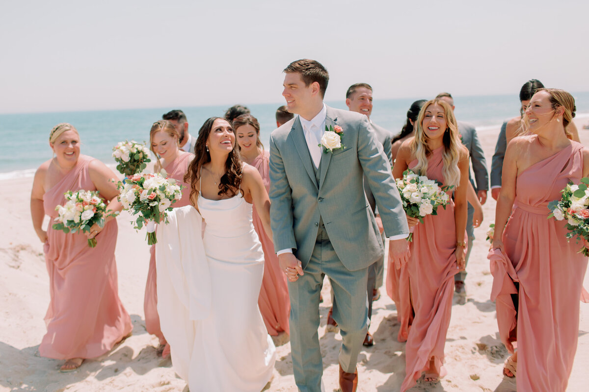 Newlyweds smiling as they walk on the beach with their wedding parties 