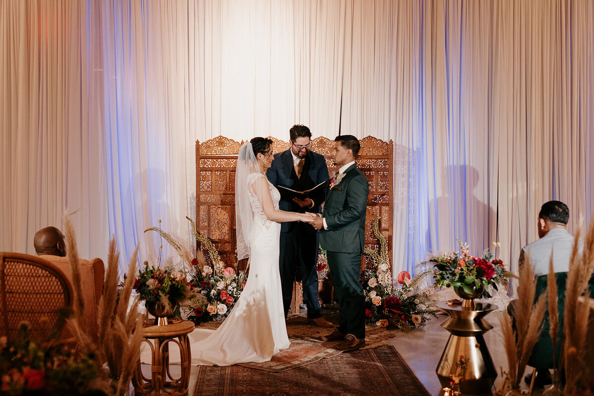 The bride and groom stand hand-in-hand at the altar, exchanging vows in front of an intricately carved wooden backdrop with vibrant florals and pampas grass surrounding them.