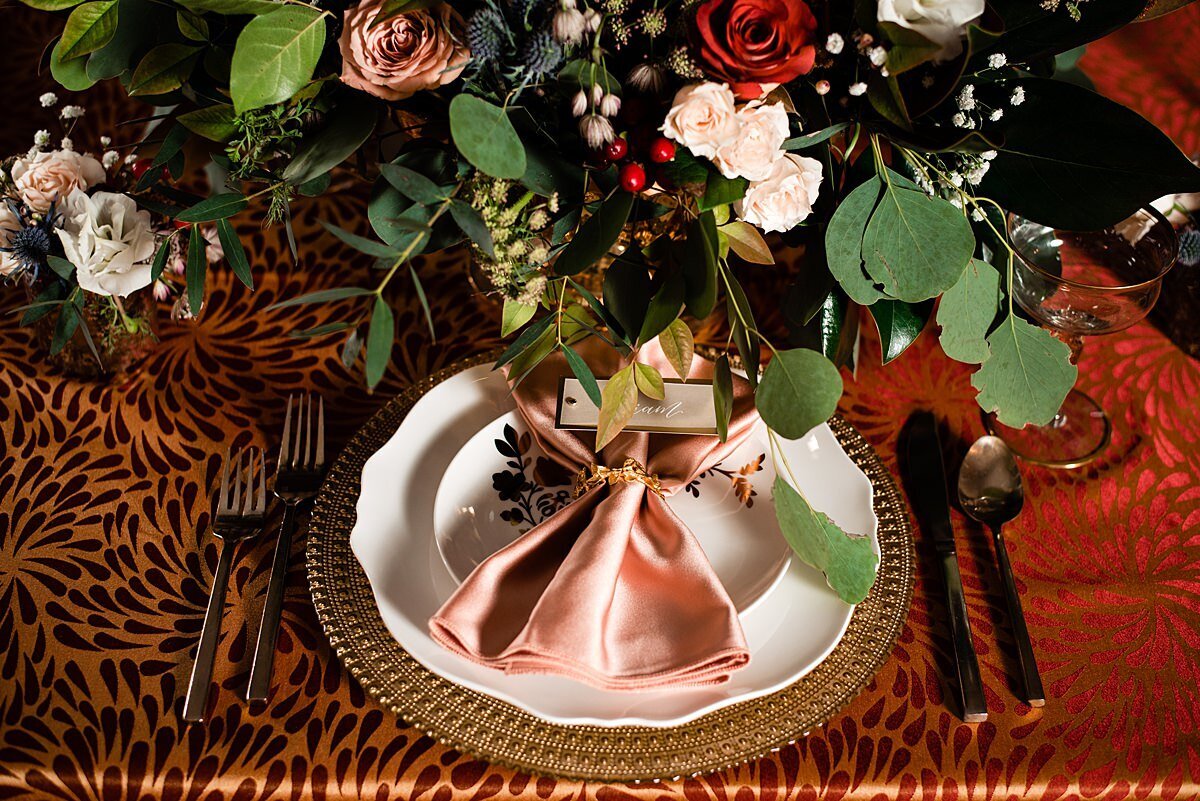 detail photo of the gold charger and white plate with the fan folded peach napkin and the large floral centerpiece with berries, peach and red roses and greenery on top of the dark orange velvet tablecloth