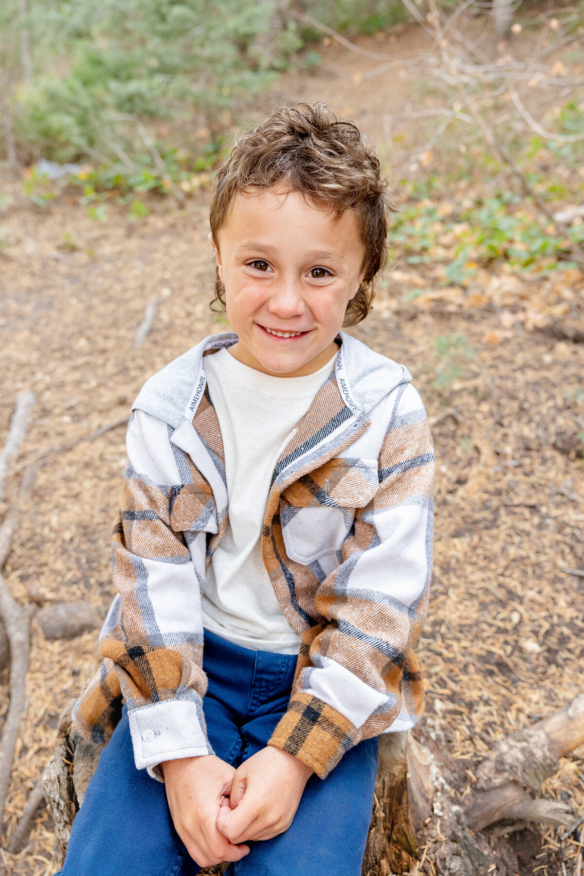A young boy sits on a tree stump, wearing a plaid flannel and blue pants, smiling warmly at the camera in a cozy, wooded environment.