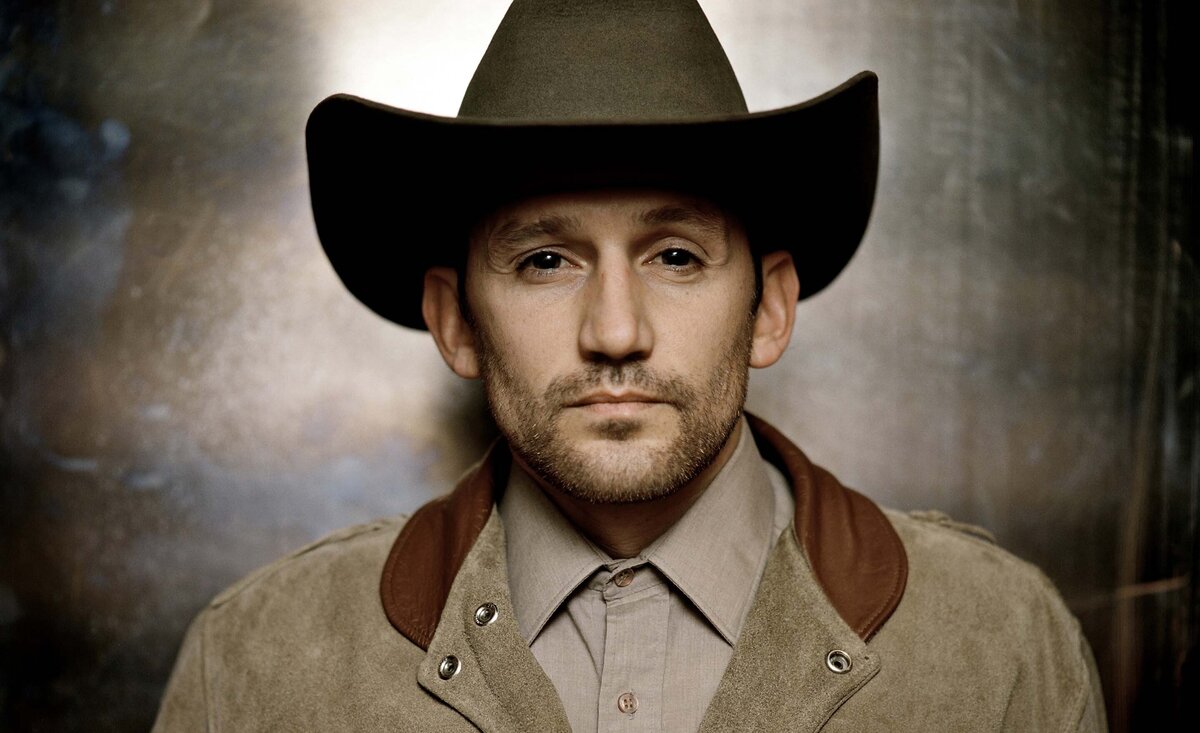 Country musician portrait Ridley Bent wearing tan coat brown cowboy hat leaning against brushed metal backdrop