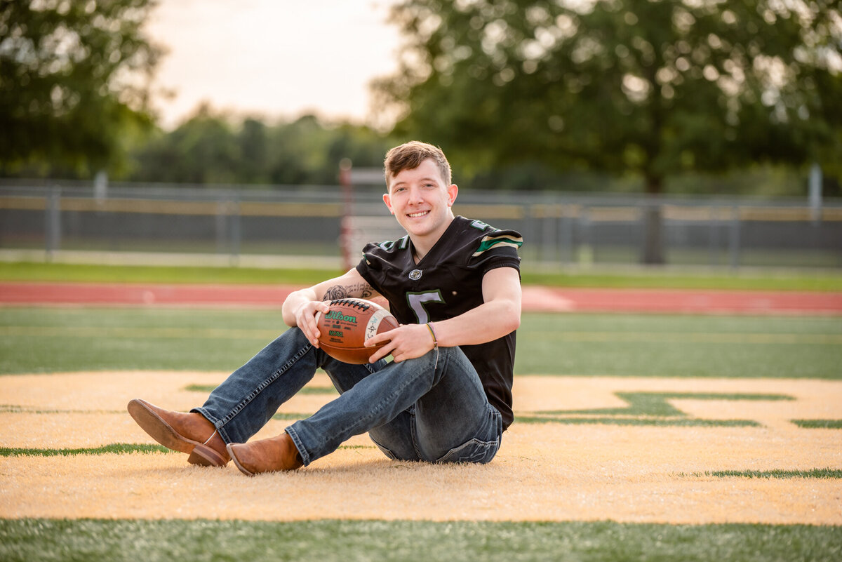 A Santa Fe High School football player sits in the end zone holding a football and wearing his jersey.