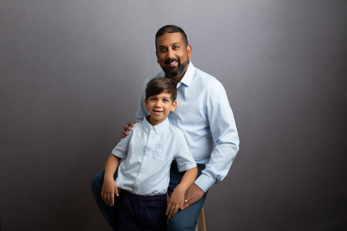 Dad sitting on a stool with his young son leaning on him and both are dressed in light blue shirts.