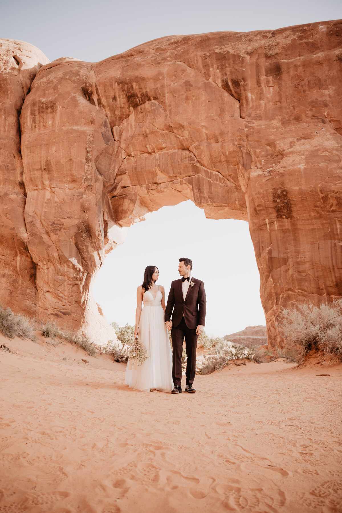 Utah elopement photographer captures bride and groom standing under arch in Arches National Park