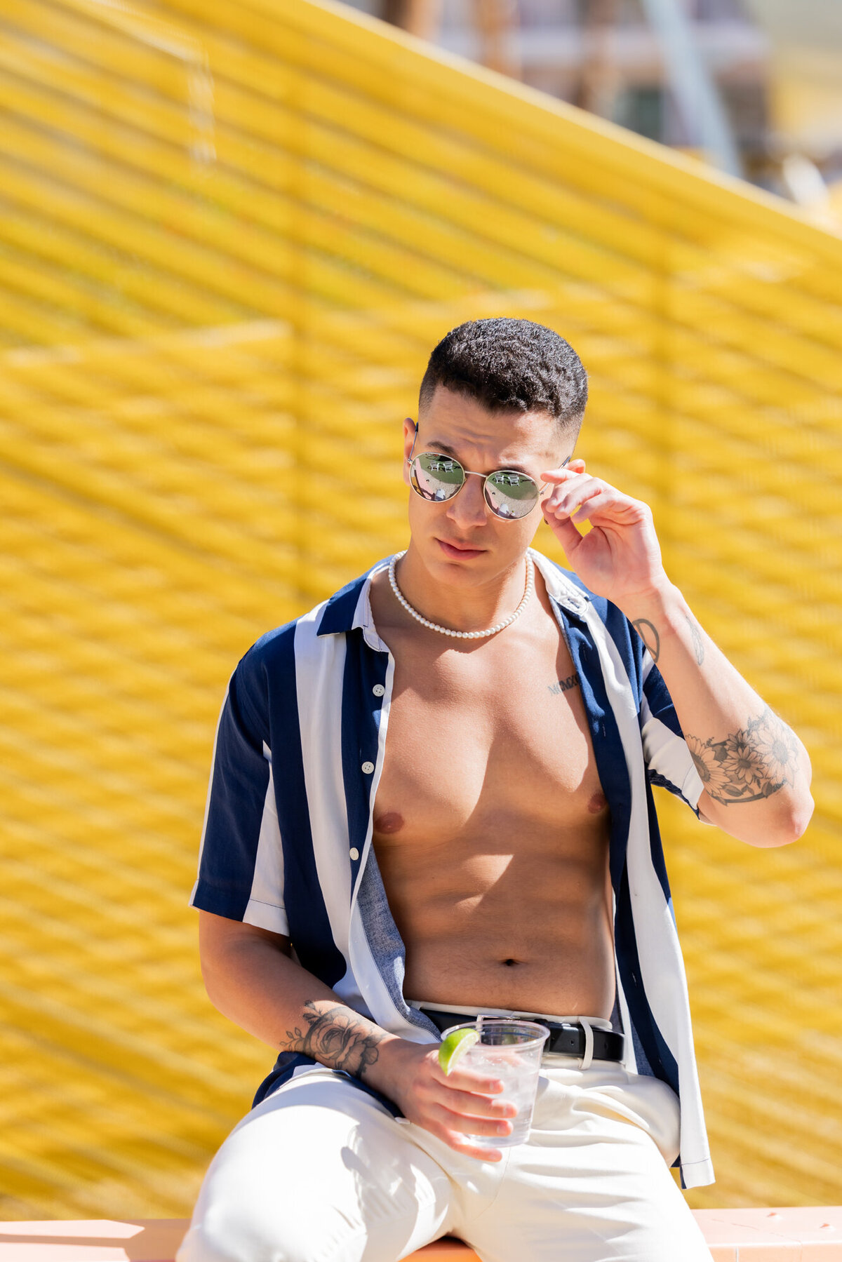 Editorial portrait of male model adjusting sunglasses against bright yellow background, styled for a poolside fashion spread.