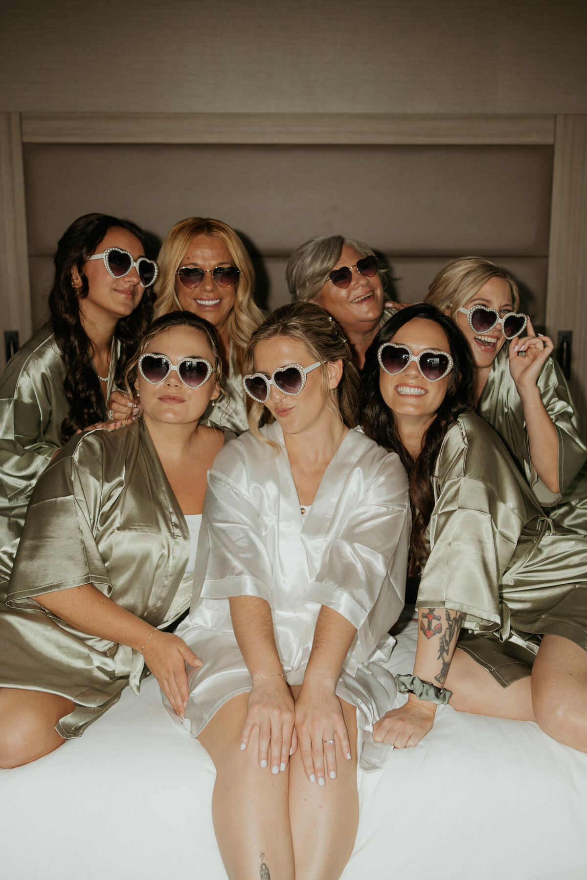 Bride and bridesmaids in matching silk robes and heart-shaped sunglasses share a candid and joyful moment on the wedding day.