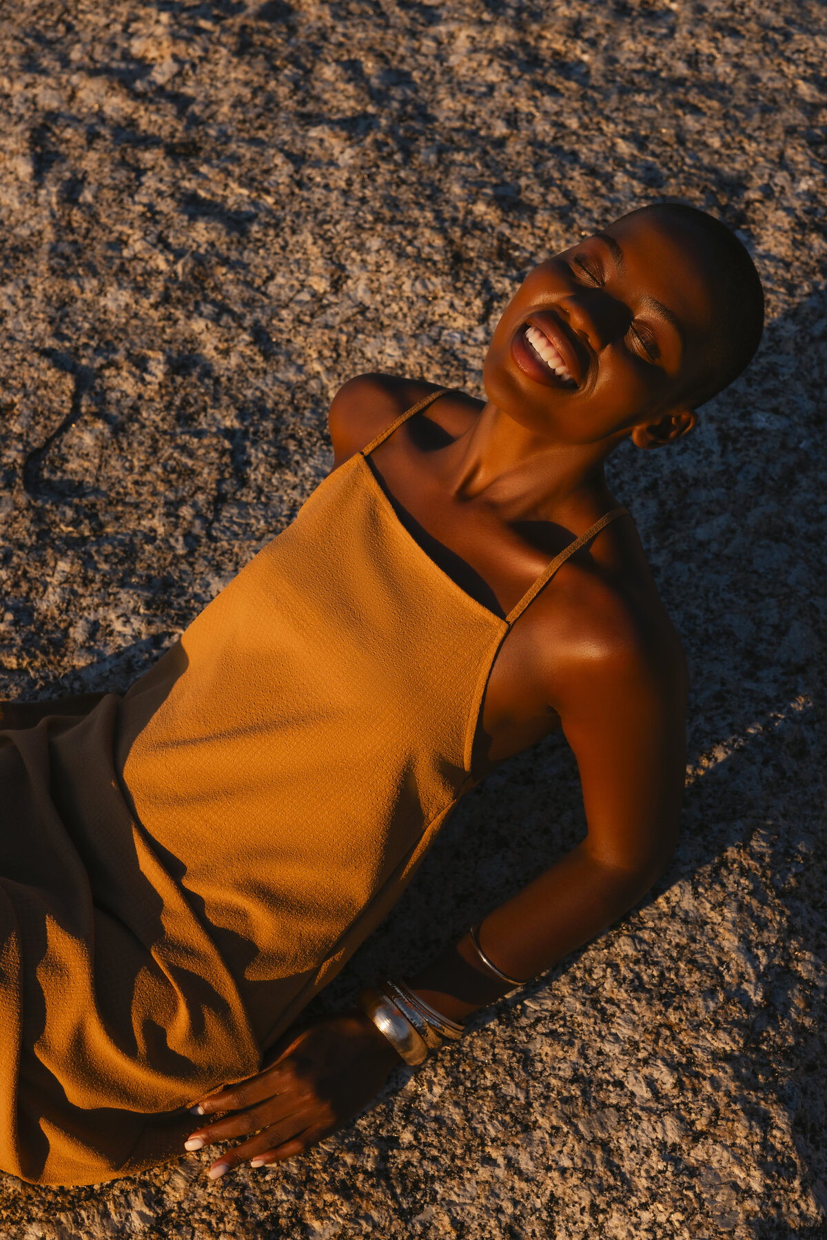 Model wearing a mustard yellow dress, smiling with eyes closed while basking in golden sunlight on textured rocks during sunset in Cape Town.