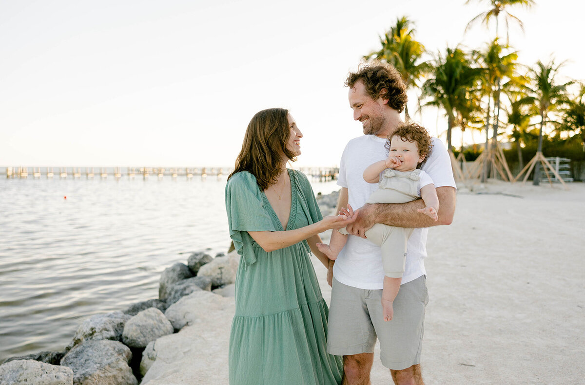  A family shares a happy moment by the ocean, with the father holding their baby while the mother looks on lovingly during a Florida Keys family session.