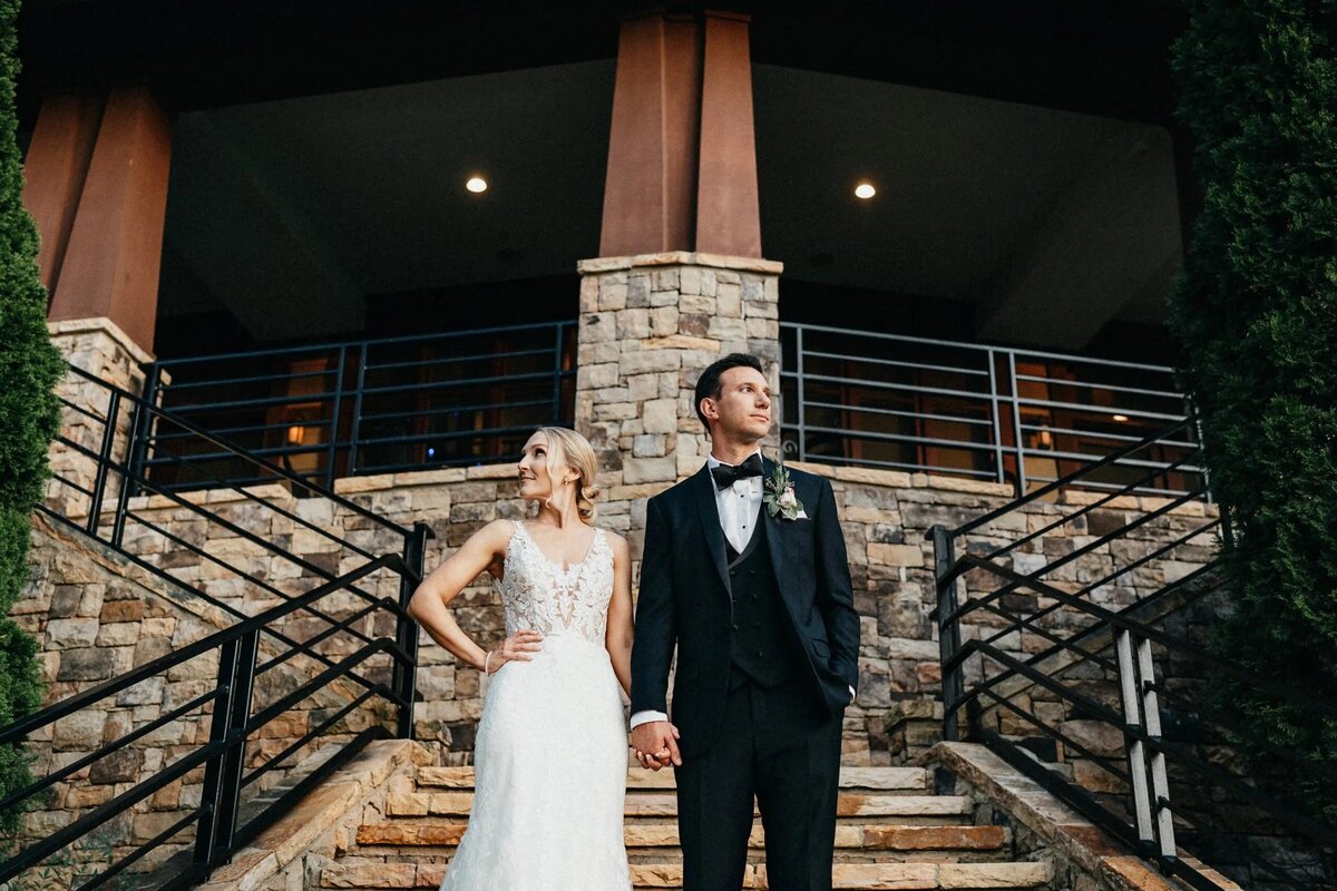 A bride and groom standing on stone steps in front of an elegant venue, looking off into the distance, evoking a sense of anticipation.