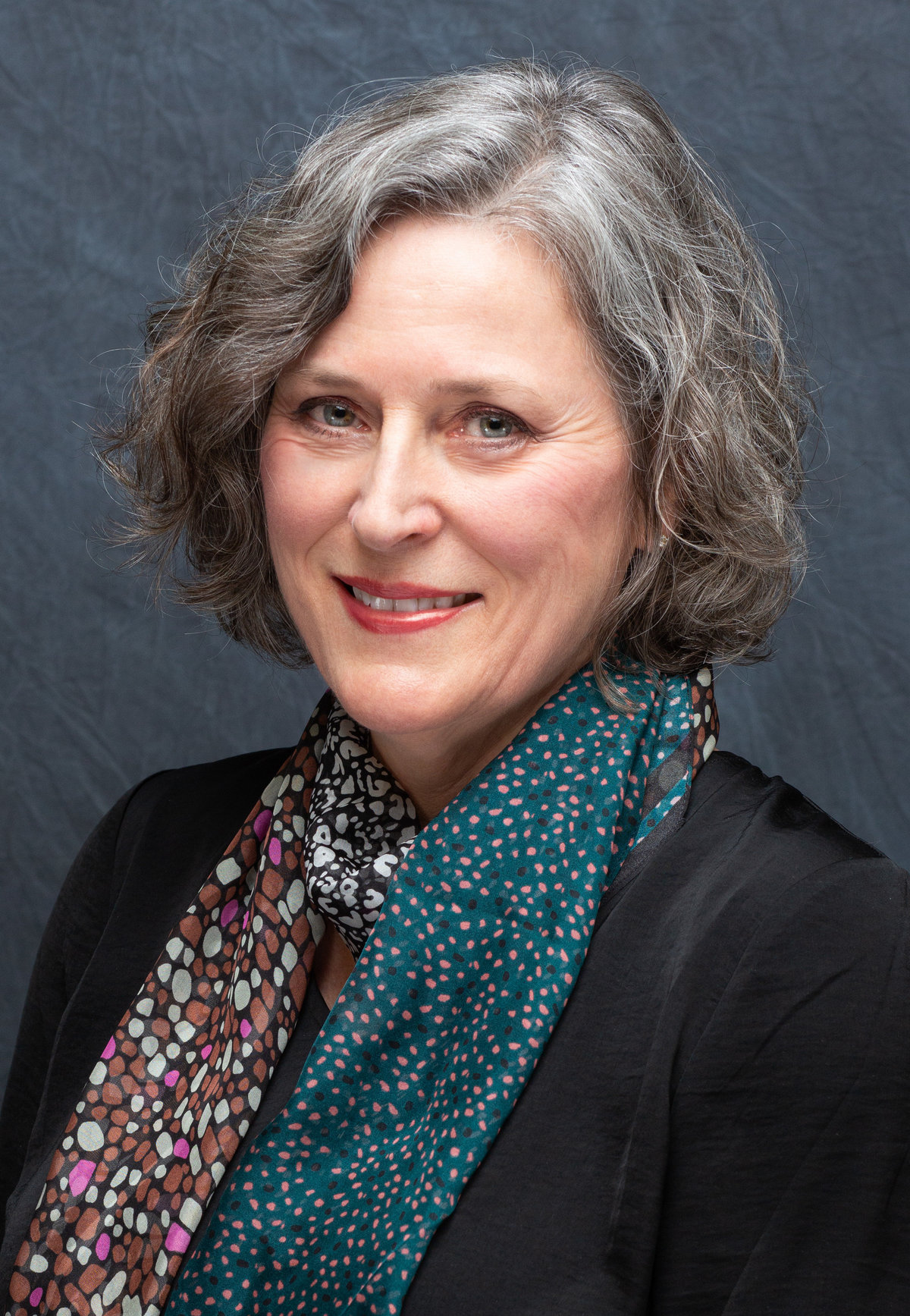 Elegant headshot portrait of a mature woman with silver hair, adorned in a multi-patterned scarf, set against a subtle gray background.