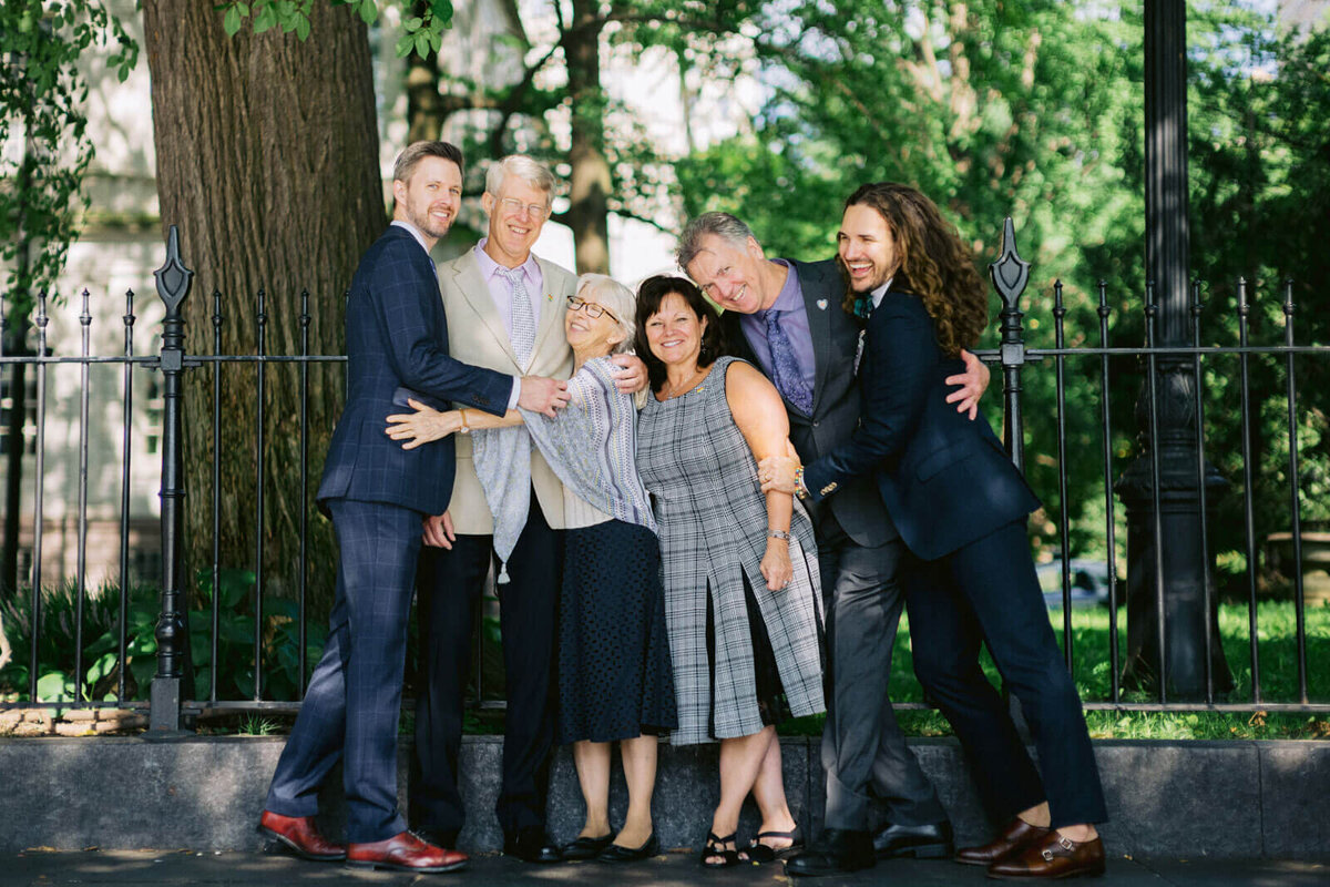 The two grooms are smiling happily with their parents. Trees in the background. NYC City Hall Elopement Image by Jenny Fu Studio
