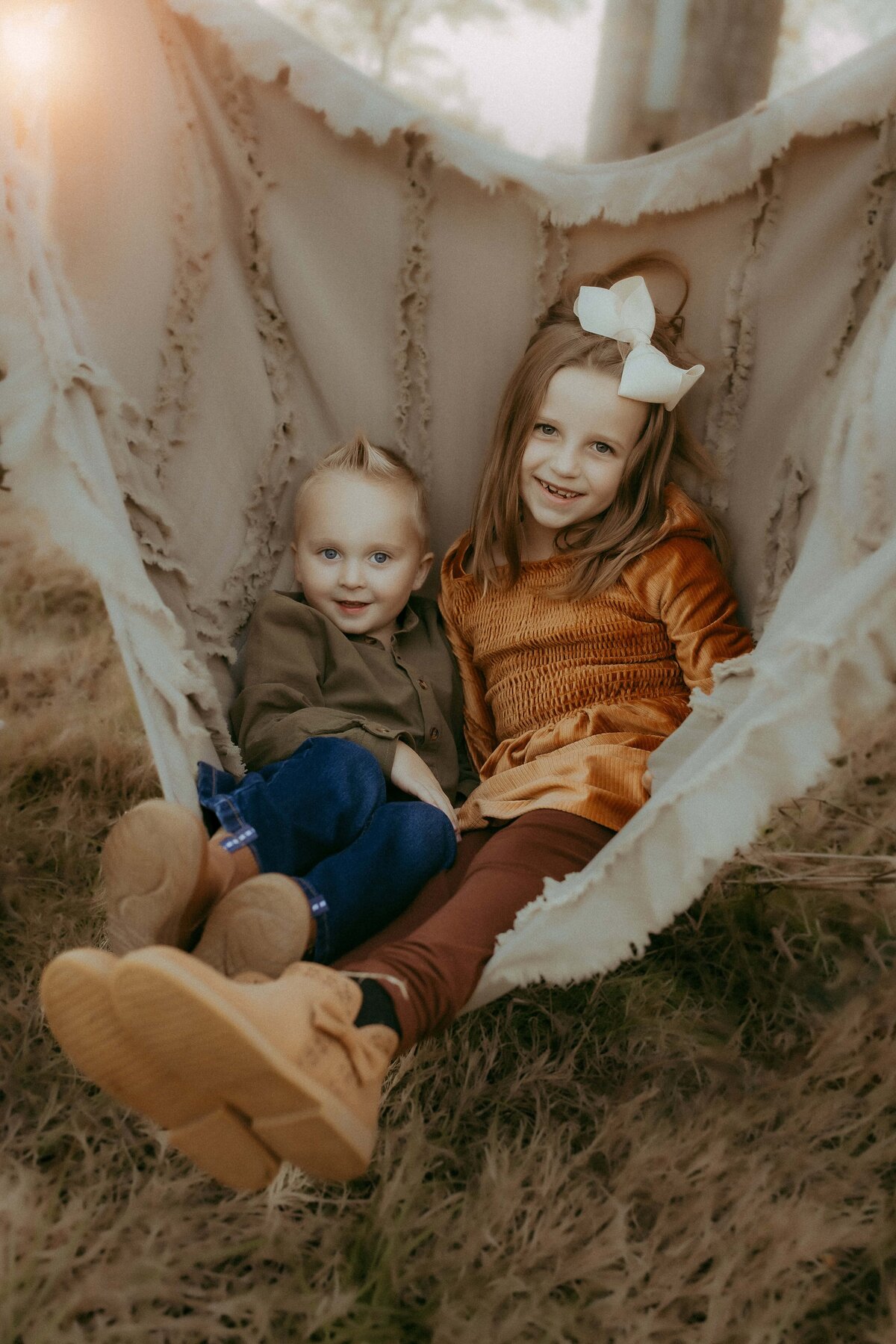 Two children, a boy and a girl, sit together on a draped hammock outdoors. The girl wears an orange dress and bow, while the boy sports a green shirt and jeans. Captured by a talented Warner Robins photographer, both children are smiling brightly.