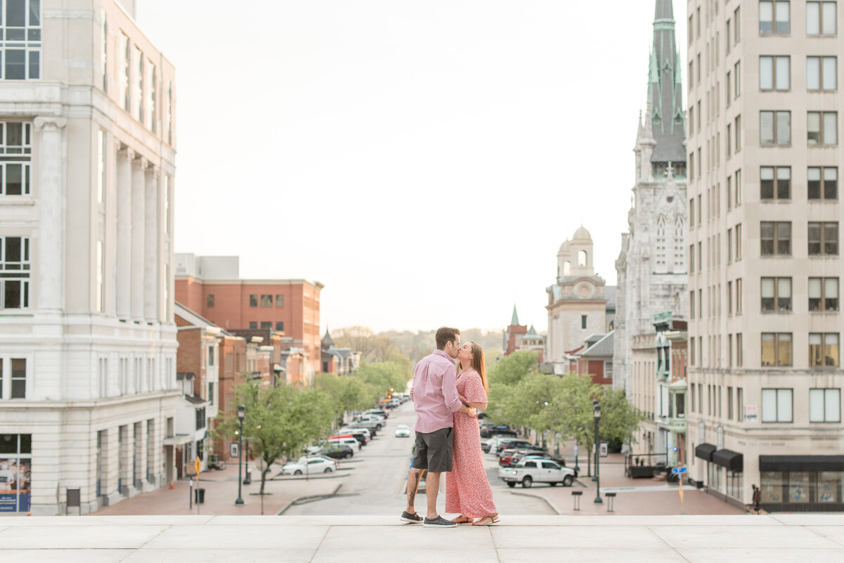 Engaged couple hugging and kissing atop steps in Harrisburg with tall city buildings beside them.