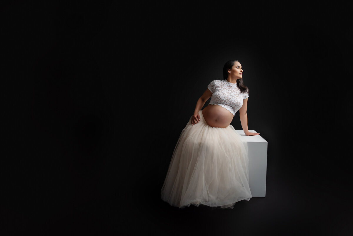 pregnant woman wearing a cream tutu skirt and white lace crop top sitting on a white box for her studio maternity photography session