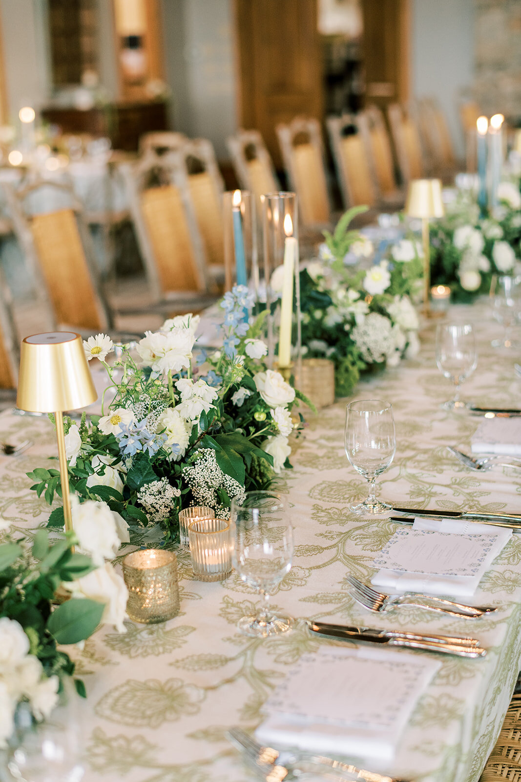  Banquet table with gold mini lamps, blue taper candles, and lush white floral greenery runner.