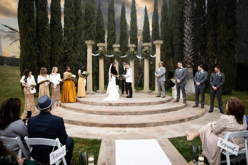 Wedding ceremony photo taken from the middle of the aisle at Grand Island mansion near sacramento. Bridesmaids in gold and groomsmen in grey look on to the bride, groom and officiant in the center.  Trees and a cloudy sky are in the background. Photo by Philippe Studio Pro from Sacramento.