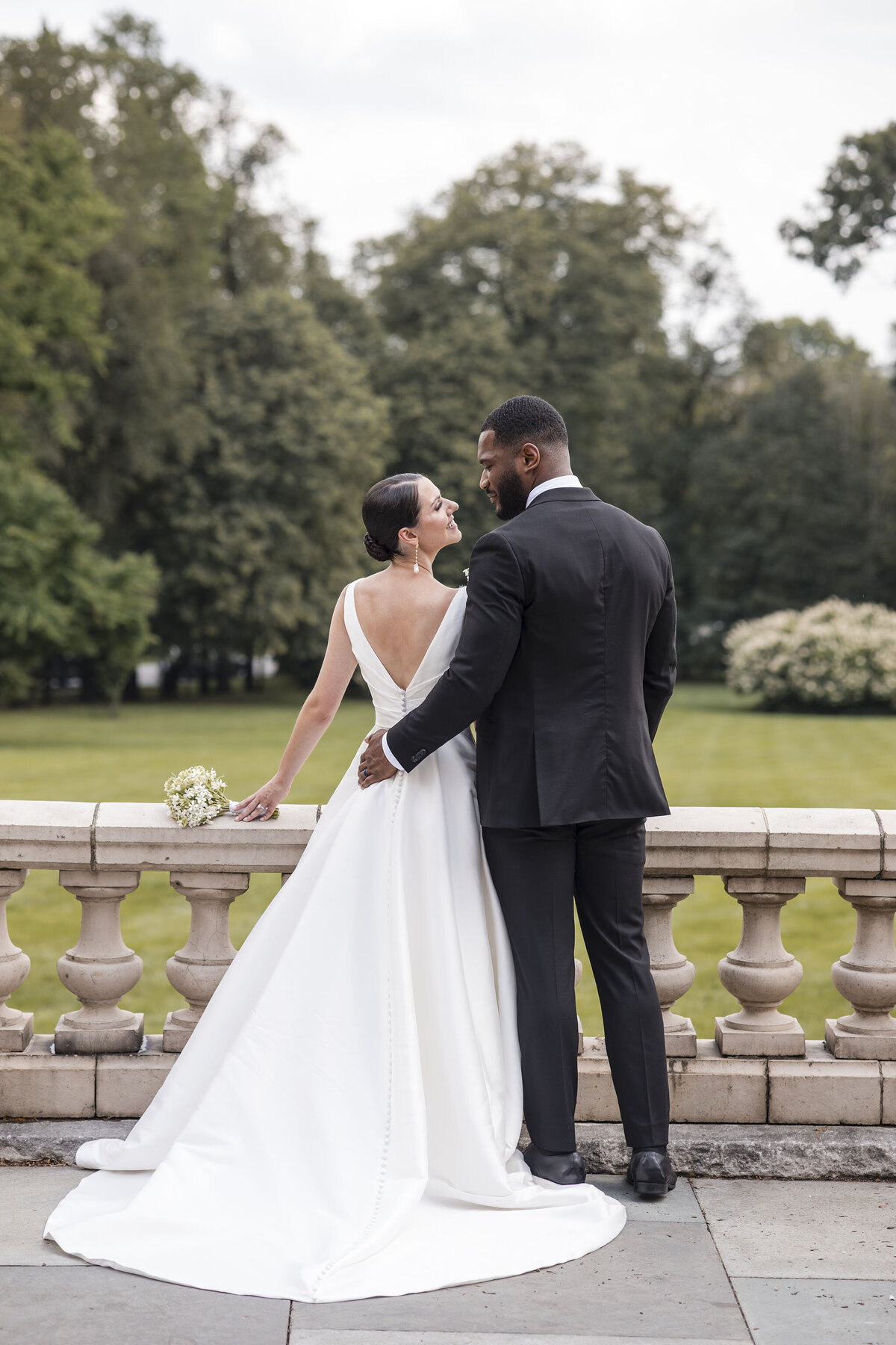 Editorial wedding portrait of a Black groom and his Caucasian bride standing in front of marble fence with an estate garden in the background.  The couple enjoys an intimate gaze, dressed in chic, elegant attire.