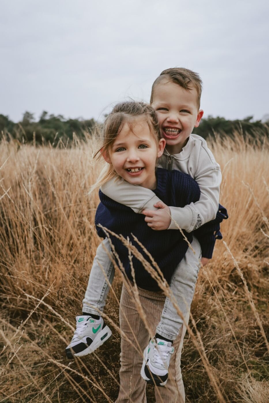 Twee lachende kinderen in een veld met lang gras, het jongetje draagt het meisje op zijn rug.