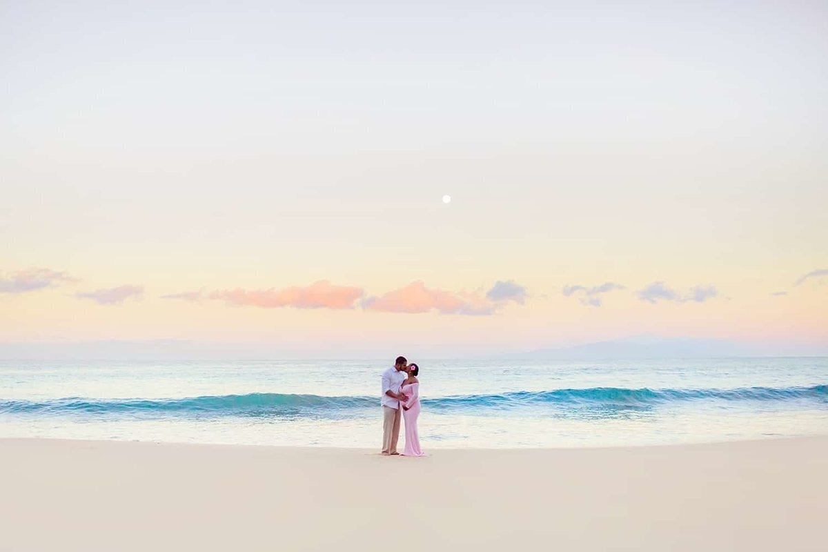 Wide portrait of a pregnant couple on Maui kissing at sunrise under beautiful pastel skies.