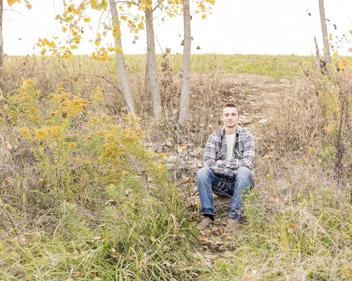 a high school senior male, sitting in the grass, wearing a flannel jacket, with a light tan shirt underneath, light brown dress shoes, blue jeans, and looking at the camera with a soft smile