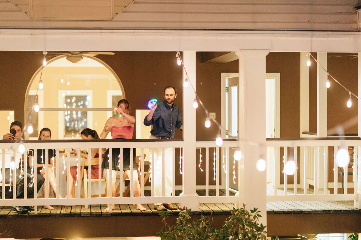 A group of people stand on a balcony decorated with string lights at night; a man holds a lit-up ball while a woman beside him blows bubbles, and others sit or take photos in a relaxed, festive atmosphere.