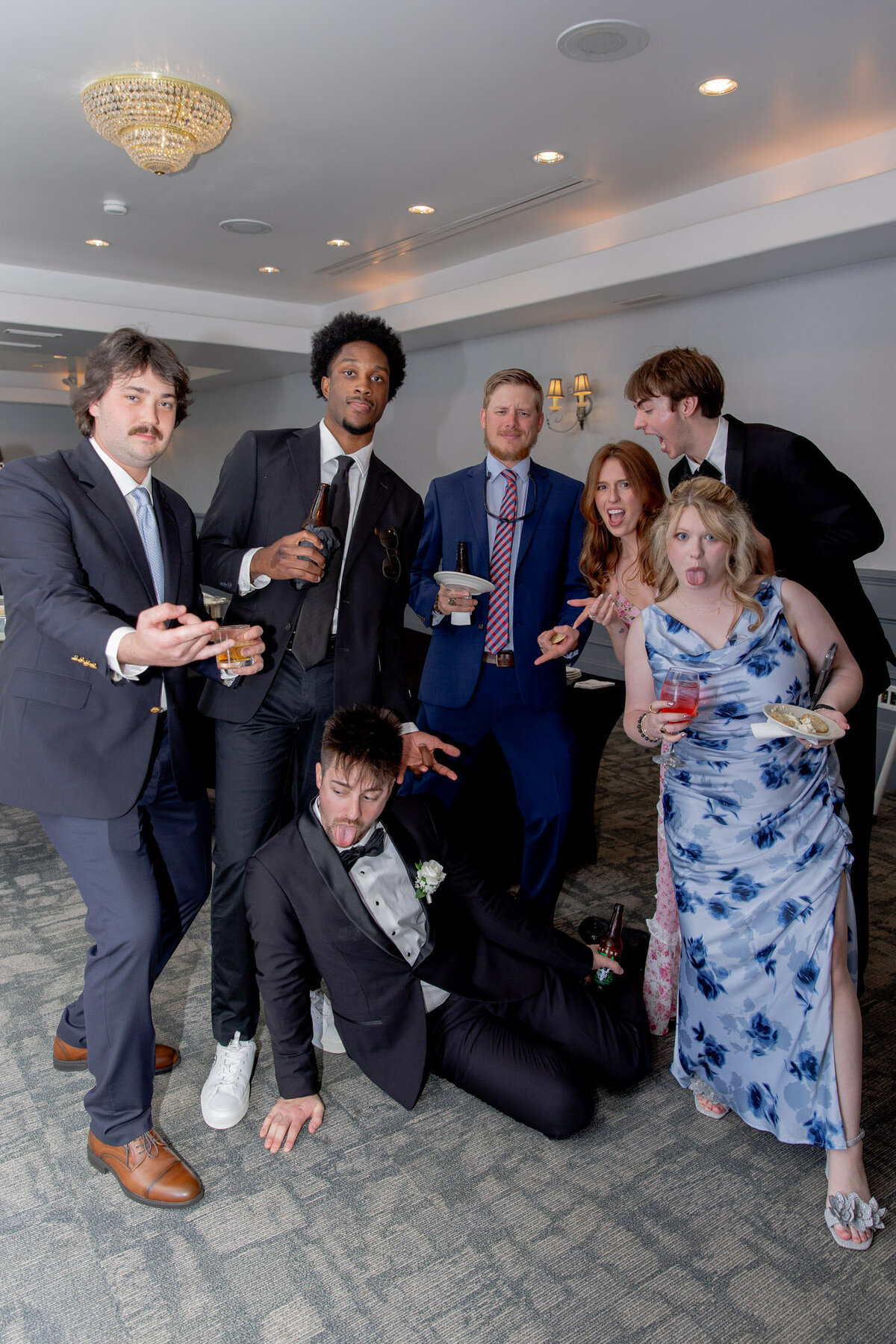 A group of men and woman pose for a silly photo, most with food or a drink in hand, during the wedding day.