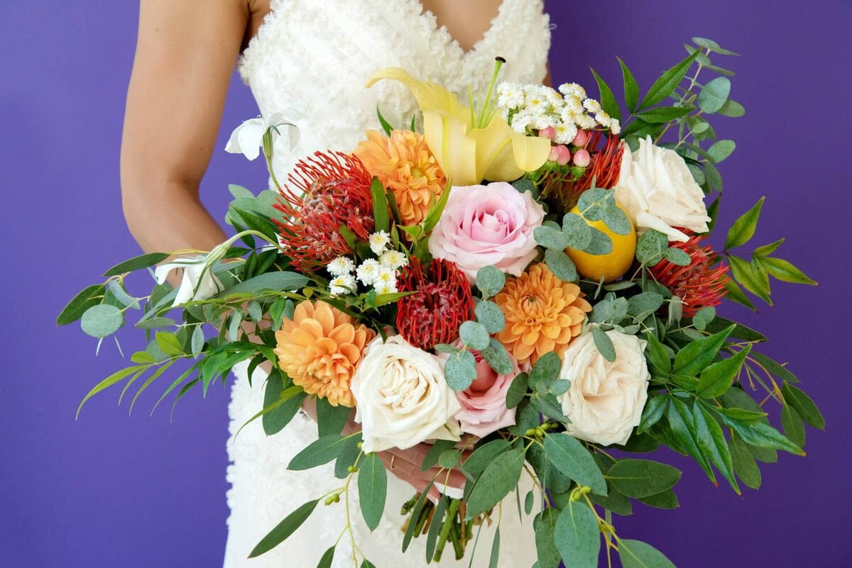 a bride holds a colorful wedding bouquet in front of a purple wall
