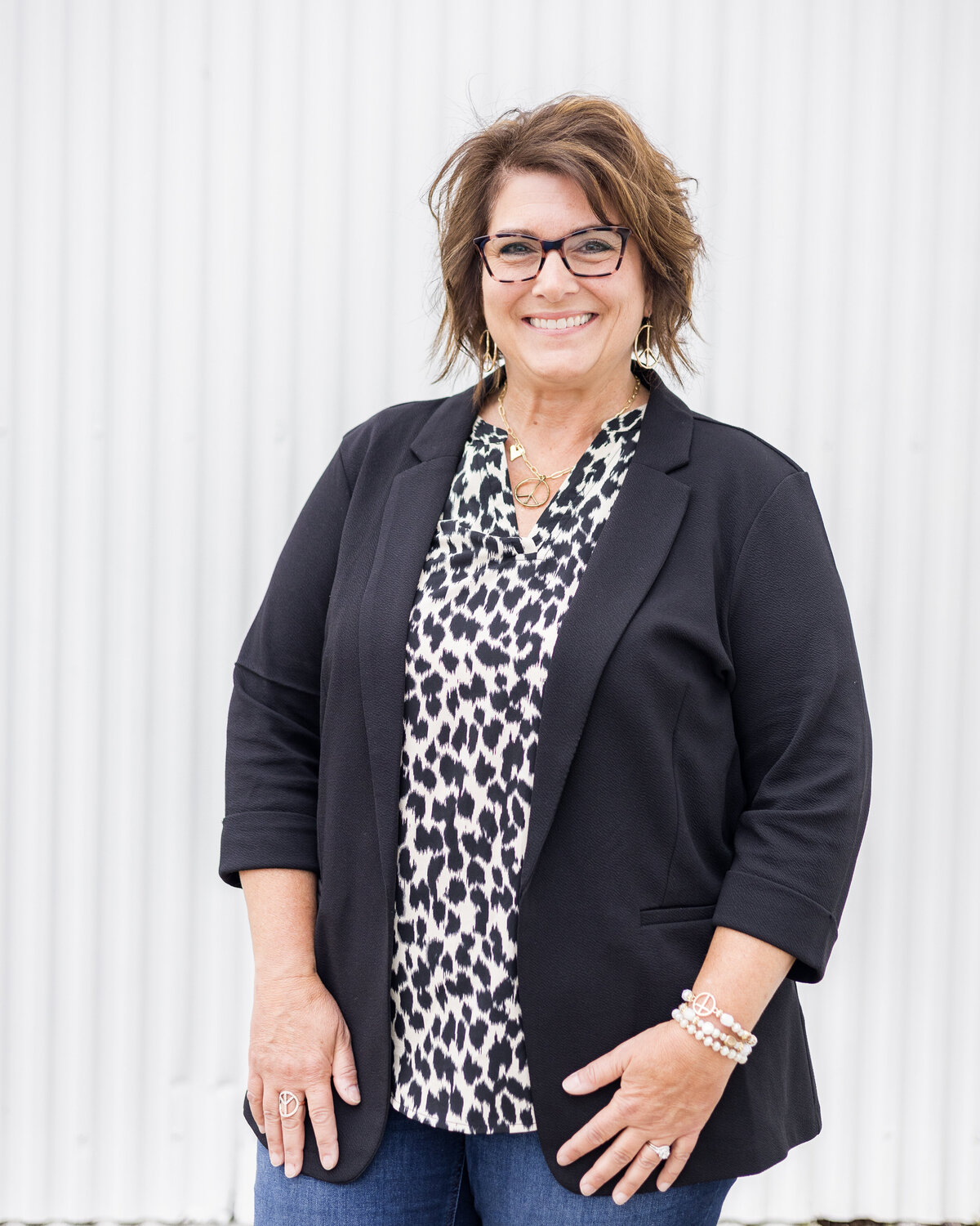 Woman standing in front of a white building with brunette blonde hair, wearing a black dress jacket with a white and black cheetah print shirt, wearing glasses, smiling at the camera