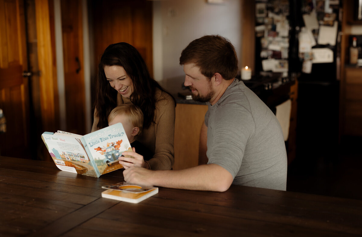 A New England Family Photographer captures a warm scene of a woman and man sitting at a table in their cozy kitchen, reading "Little Blue Truck" to a small child. The homey atmosphere is accented by a fridge adorned with magnets and cherished pictures in the background.