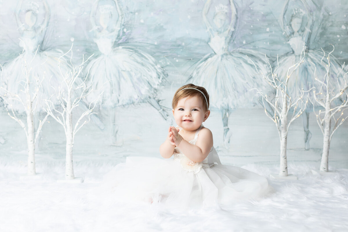 Dallas milestone photographer captures baby girl in a white gown sitting on soft white fur with whimsical ballerina backdrop and twinkling birch trees.
