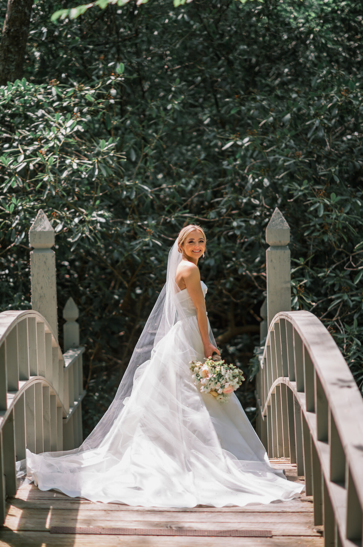 Bride in a strapless white gown and long veil standing on a wooden bridge surrounded by greenery at Old Edwards Inn in Highlands, North Carolina, holding a pastel bouquet.