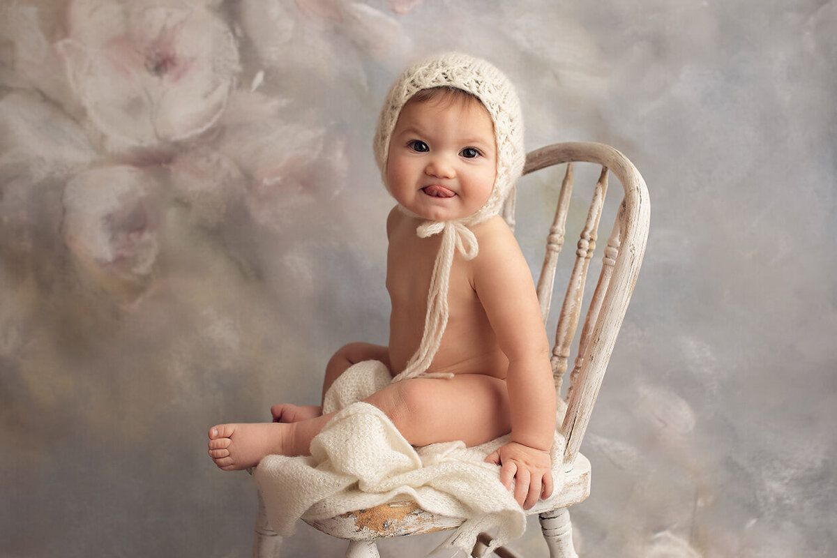 a one year old sitting in a white chair wearing a white bonnet with her tongue sticking out