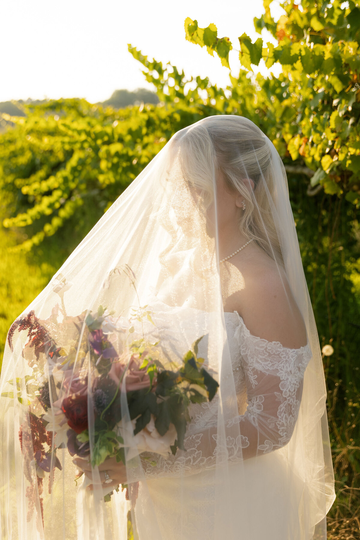 A bridal portrait in a sunlit vineyard with the bride standing beneath a soft, flowing veil while holding a textured floral bouquet filled with mauve roses, deep red blooms, trailing amaranthus, and organic greenery, designed by a wedding florist for an outdoor ceremony.