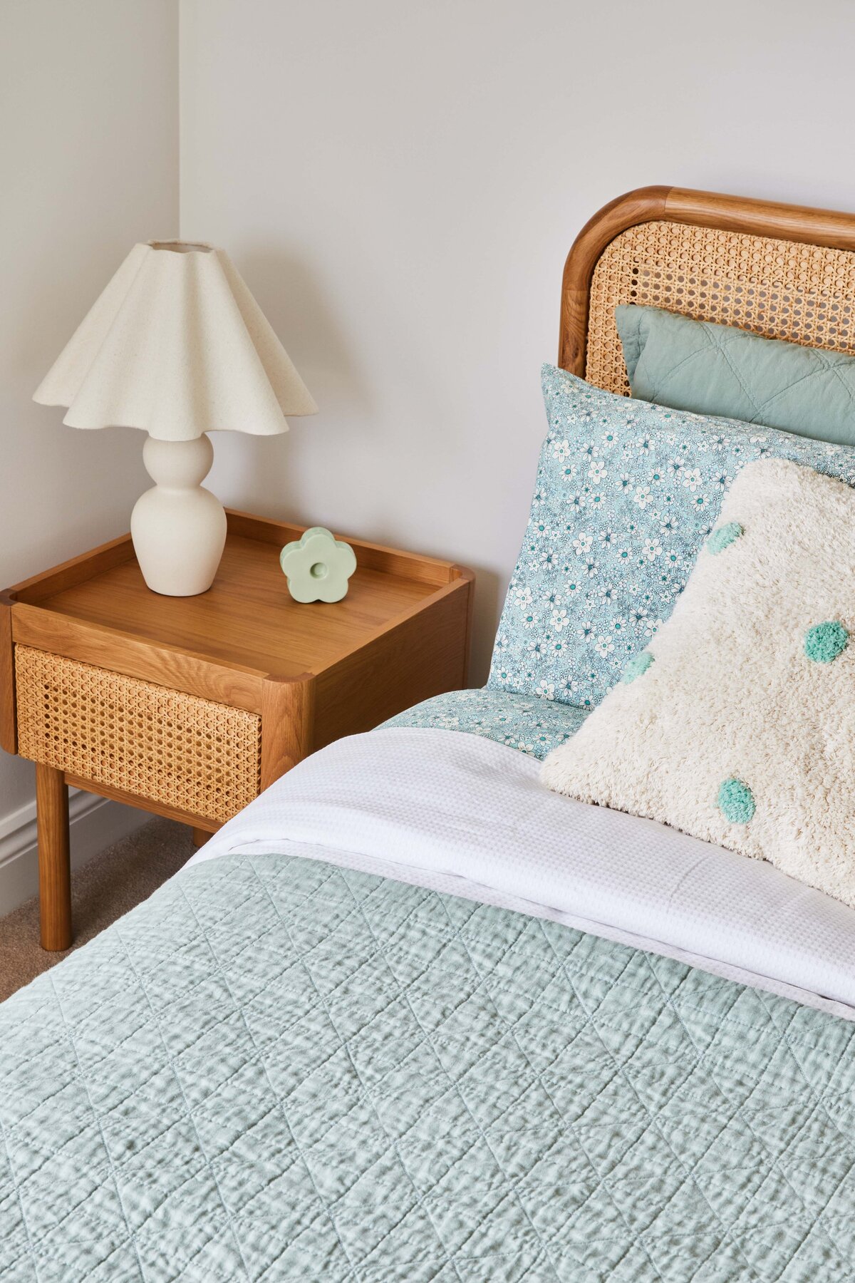 Stylish bedroom. The wooden table has a cane front and is styled with a contemporary lamp and a small green ceramic ornament. The bed features a light green quilted coverlet and a cane headboard.