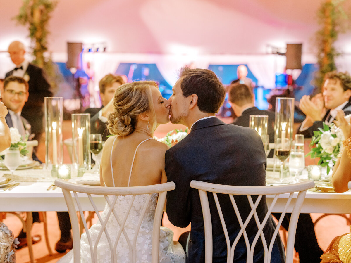 Photo from behind of bride and groom kissing while sitting at their reception table