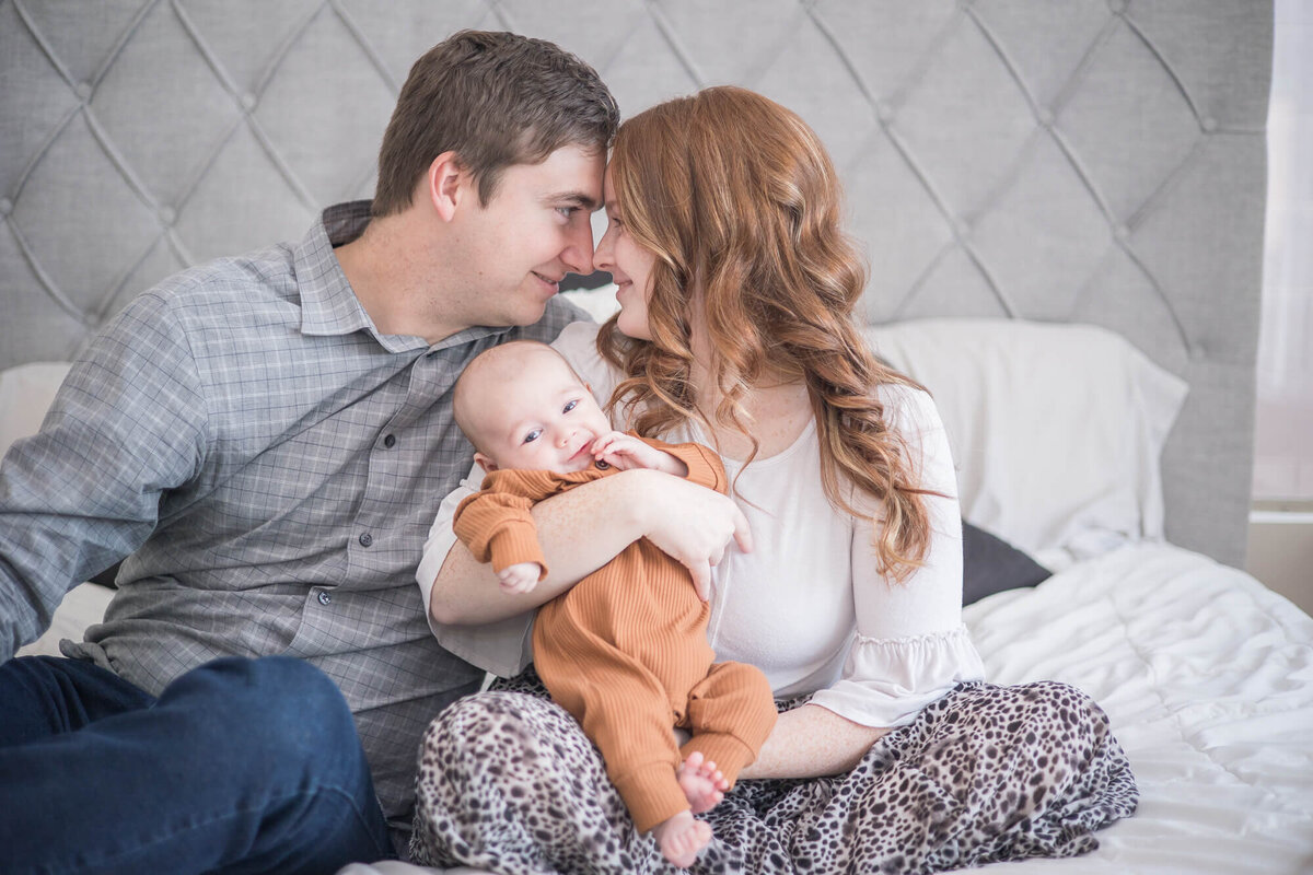 Couple cheekily nuzzling noses while holding infant son on white and gray bed