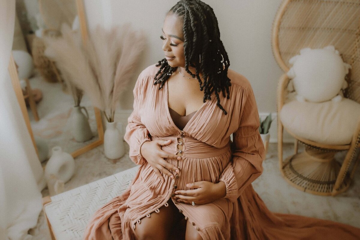 Pregnant woman with braided hair and a long peach dress sits touching her belly in a cozy room with pampas grass and wicker furniture.