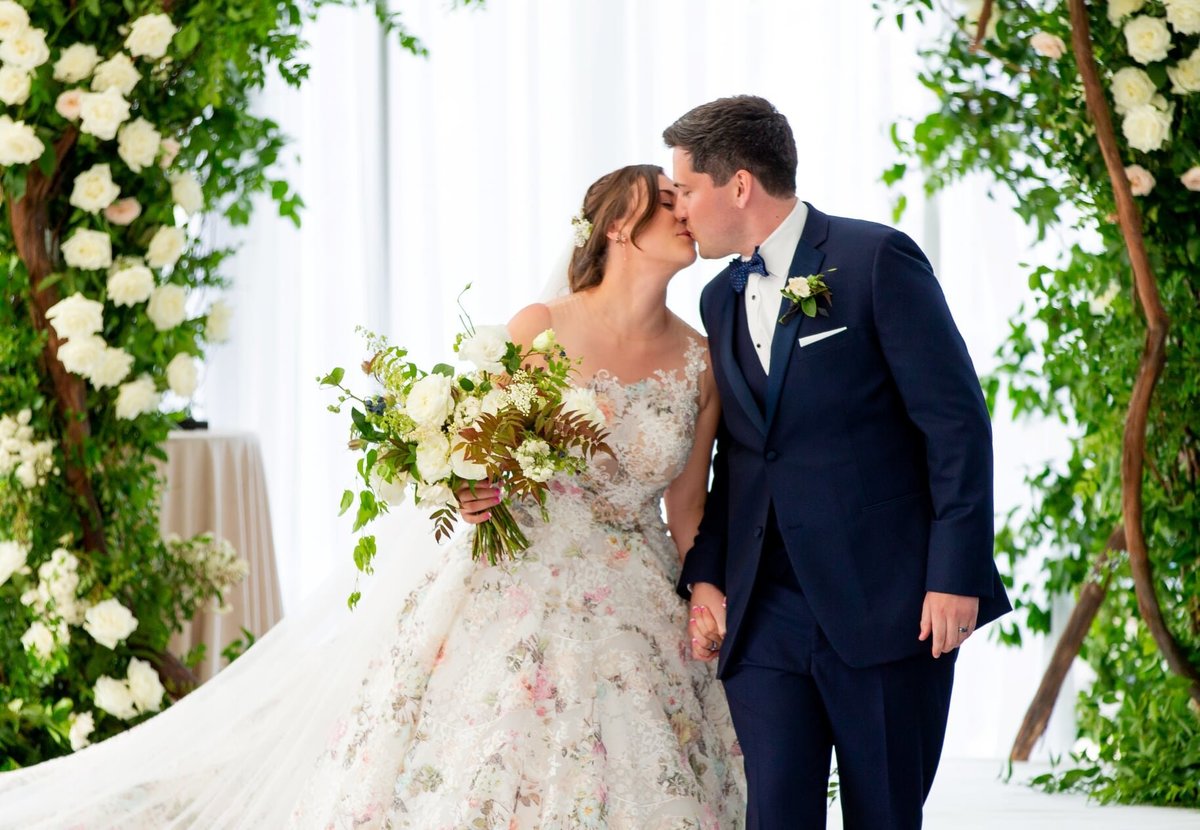 Newly wed couple kissing in front of grand greenery wedding arbor
